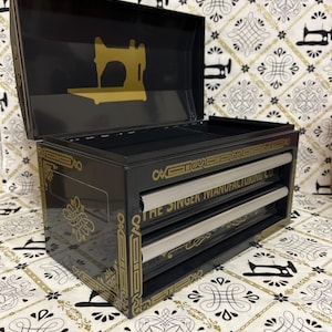 May include: Black sewing machine storage box with gold accents. The box features a gold sewing machine silhouette on the lid and the text "THE SINGER MANUFACTURING CO." on the front. The box has three drawers.