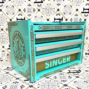 May include: A turquoise metal storage cabinet with three drawers, featuring gold decorative borders and the Singer Manufacturing Co. logo on the side. The bottom drawer displays the word SINGER in large gold letters, while the middle drawer reads The Singer Manufacturing Co. in gold text.