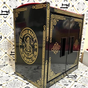 May include: A black Singer Manufacturing Co. cabinet with gold accents and a red top. The cabinet features the Singer logo and decorative gold trim. The cabinet has two doors and is likely used for storing sewing supplies.