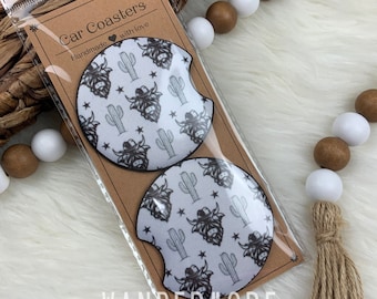 Puede incluir: Dos posavasos de coche blancos con un patrón en blanco y negro de una vaca de las tierras altas con un sombrero de vaquero y cactus. Los posavasos están empaquetados en una funda de plástico transparente con un respaldo de cartón marrón que dice "Car Coasters Handmade with love" y "Wandermore".