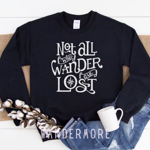 以下が含まれることがあります： 白い文字で「Not all who wander are lost」と書いてある黒いスウェットシャツ。コンパスのデザインが施されています。スウェットシャツは白い表面に置かれ、青いジーンズと白いマグカップが置かれています。