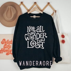 以下が含まれることがあります： 黒いスウェットシャツに、白い文字で「Not all who wander are lost」と書いてあり、コンパスのグラフィックが描かれています。スウェットシャツの下部に白い文字で「WANDERMORE」と印刷されています。