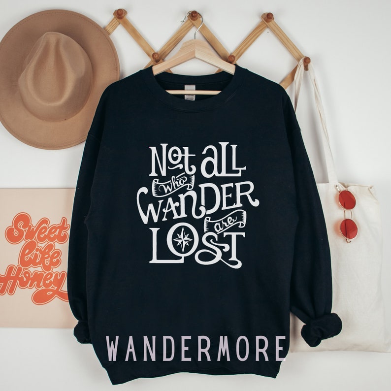 以下が含まれることがあります： 黒いスウェットシャツに、白い文字で「Not all who wander are lost」と書いてあり、コンパスのグラフィックが描かれています。スウェットシャツの下部に白い文字で「WANDERMORE」と印刷されています。