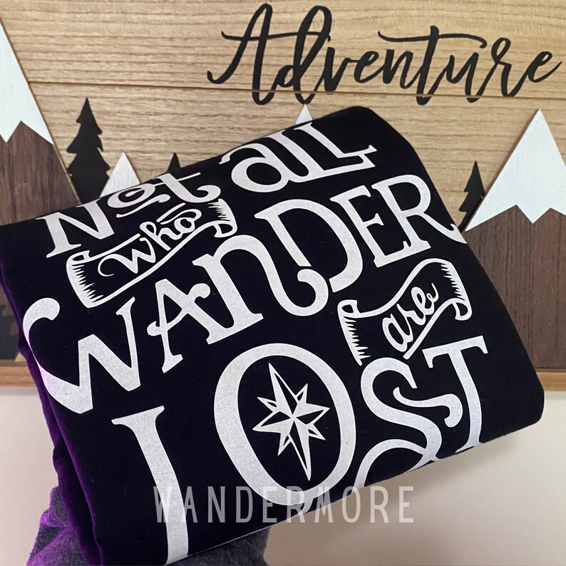 以下が含まれることがあります： 黒いスウェットシャツに、白い文字で「Not all who wander are lost」と、コンパスのグラフィックが描かれています。グラフィックの下には「Wandermore」という文字が印刷されています。