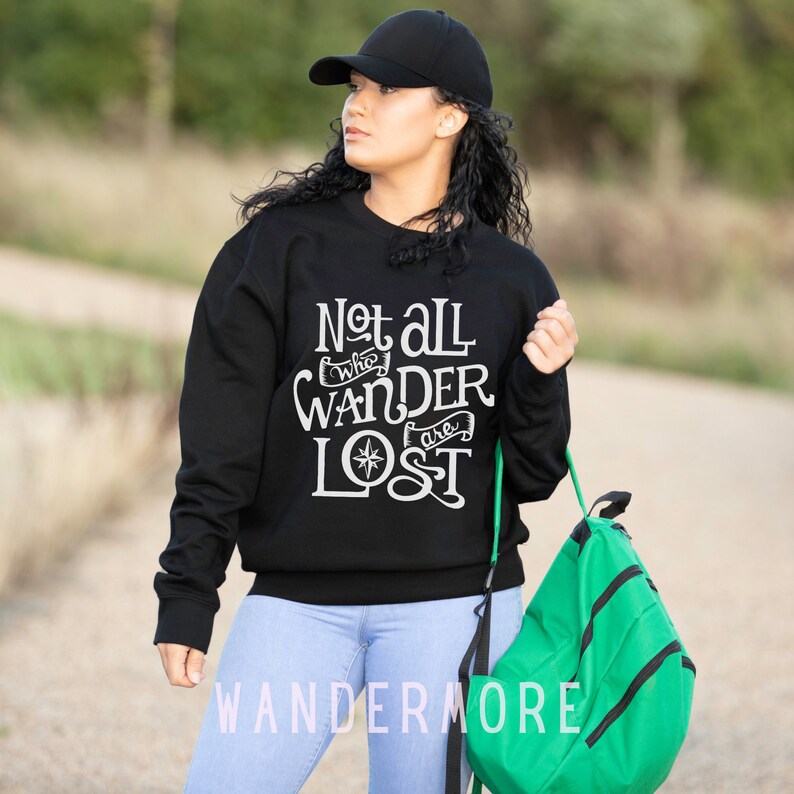 以下が含まれることがあります： 黒いスウェットシャツに、白い文字で「Not all who wander are lost」と書いてあり、コンパスのグラフィックが描かれています。モデルは青いジーンズと黒いベースボールキャップを着用しています。