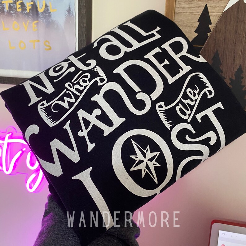 以下が含まれることがあります： 黒いTシャツに白い文字で「Not all who wander are lost」とコンパスのグラフィックが書かれています。グラフィックの下に「Wandermore」という文字が印刷されています。