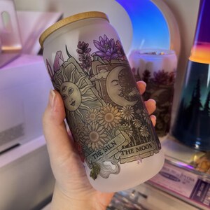 May include: Frosted glass can-shaped tumbler with a bamboo lid. The design features a sun with three eyes, a crescent moon, flowers, and the text "THE SUN THE MOON". The tumbler is held in a person's hand.