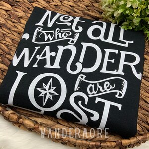 以下が含まれることがあります： 黒いTシャツに白い文字で「Not all who wander are lost」とコンパスのグラフィックが書かれています。グラフィックの下には「Wandermore」という文字が印刷されています。