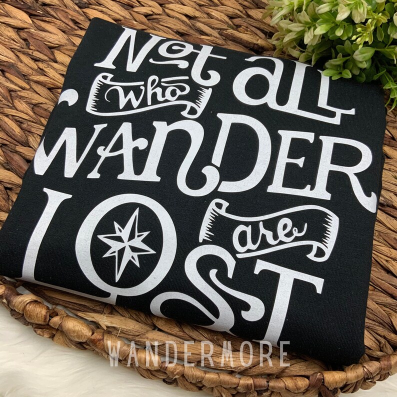 以下が含まれることがあります： 黒いTシャツに白い文字で「Not all who wander are lost」とコンパスのグラフィックが書かれています。グラフィックの下には「Wandermore」という文字が印刷されています。