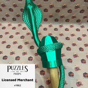 May include: A green 3D printed cobra snake staff topper with a crystal-shaped head. The staff topper is on a wooden staff. The staff topper is labeled "Puzzles and Props Licensed Merchant #1962".