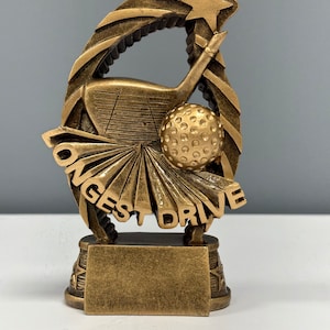May include: Gold-colored golf trophy with a golf club and ball design. The trophy has a star at the top and the words "Longest Drive" on the base.