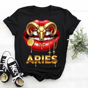 Puede incluir: Camiseta negra con un diseño de cabeza de carnero dorada sobre labios rojos. La palabra "ARIES" está en oro, con letras goteando. Zapatillas deportivas negras y blancas en la parte inferior.