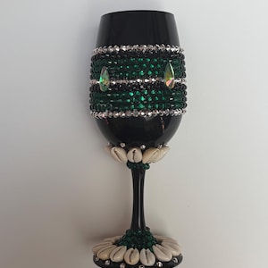 May include: A black wine glass with decorative embellishments. The glass features a band of green and silver beads, accented with iridescent teardrop-shaped gems. The base is adorned with seashells and green beads.