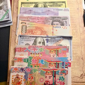 May include: A collection of various international paper currency replicas and ancestor money notes arranged on a clipboard. The stack includes bills with portraits, bridge illustrations, and Chinese characters in vibrant colors like pink, blue, and orange.