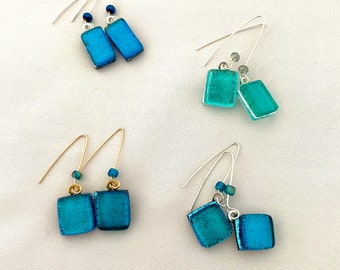 May include: Four pairs of dangle earrings with square glass beads in shades of blue and teal. The earrings are on a white surface.