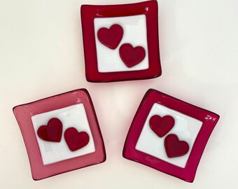May include: Three small square glass dishes with red and pink borders. Each dish has a white centre with two red hearts.