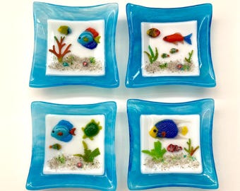 May include: Set of four square glass dishes with blue borders. Each dish features a different colourful glass mosaic of fish and sea life on a white background.