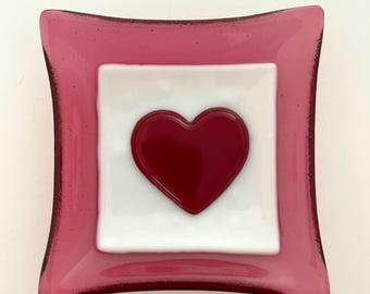 May include: A small, square glass dish with a pink border and a white centre. A red heart is in the centre of the white square.