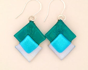 May include: Teal and blue glass earrings with a geometric diamond shape. The earrings are made of two layers of glass, with the top layer being a darker teal and the bottom layer being a lighter blue. The earrings are suspended from silver hooks.