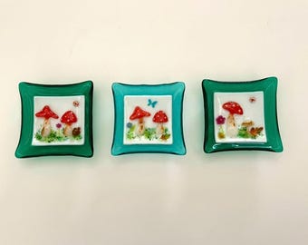 May include: Three small square teal glass dishes with white interiors and a mushroom design. Each dish features red and white mushrooms with green foliage and a butterfly on one dish.