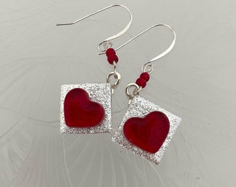 May include: Silver earrings with red glass heart charms. The hearts are set in square frames with a textured finish.