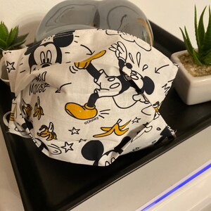 Op de afbeelding: Een wit mondkapje met een zwart en geel Mickey Mouse-ontwerp. Het mondkapje heeft de tekst "Mickey Mouse" en "©Disney" erop gedrukt.