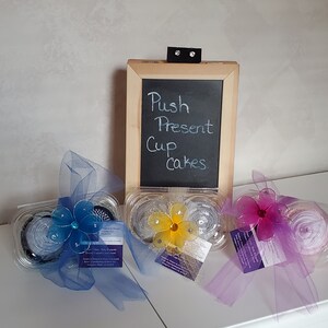 May include: Three cupcake containers, each with a sheer ribbon and a decorative flower. A small chalkboard sign reads "Push Present Cupcakes". The cupcakes are decorated with frosting. The image is of a food item.