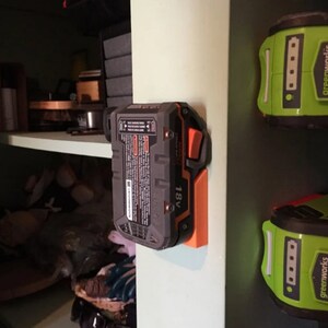 May include: A black and orange 18V battery is mounted on a white wall with an orange plastic holder. The battery has a black button on the side.