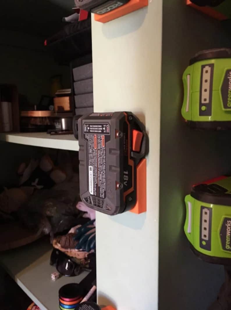 May include: A black and orange 18V battery is mounted on a white wall with an orange plastic holder. The battery has a black button on the side.
