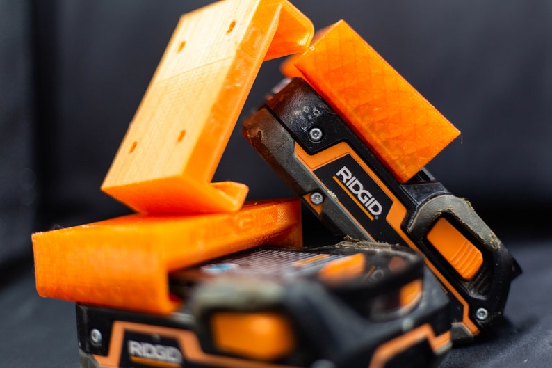 May include: Two orange 3D printed battery holders with a black and orange Ridgid power tool battery.