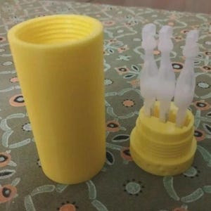 Puede incluir: Un recipiente de plástico amarillo con una tapa a juego y varios aplicadores transparentes. El recipiente es cilíndrico y la tapa tiene un diseño escalonado. Los aplicadores están dispuestos en un patrón circular.