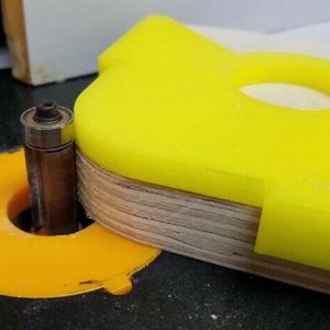May include: A yellow plastic router jig with a hole in the center, used for woodworking projects. The jig is attached to a piece of plywood and is being used with a router bit.