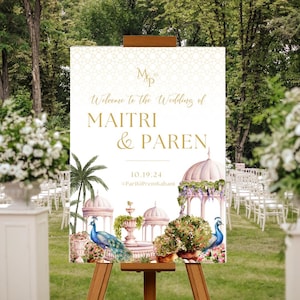 May include: A wedding welcome sign with a white background and gold lettering. The sign features a watercolor illustration of a pink and white palace with peacocks and palm trees. The text on the sign reads "Welcome to the Wedding of Maitri & Paren 10.19.24 #ParikiiPremkahaani".