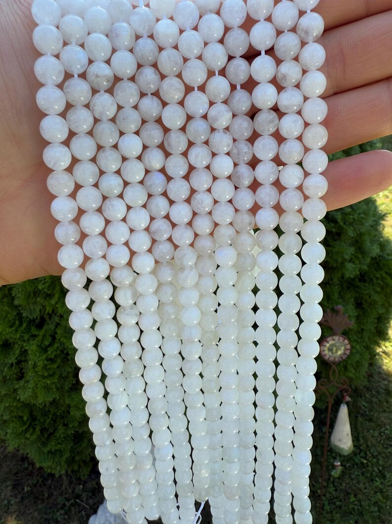 May include: Strands of white, round gemstone beads. The beads have a smooth, polished surface and are strung together on a white cord. The beads are a milky white color with subtle variations in tone. The background is a blurred green.