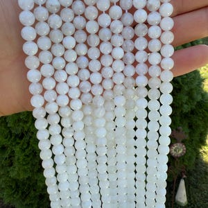 May include: Strands of white, round gemstone beads. The beads have a smooth, polished surface and are strung together on a white cord. The beads are a milky white color with subtle variations in tone. The background is a blurred green.