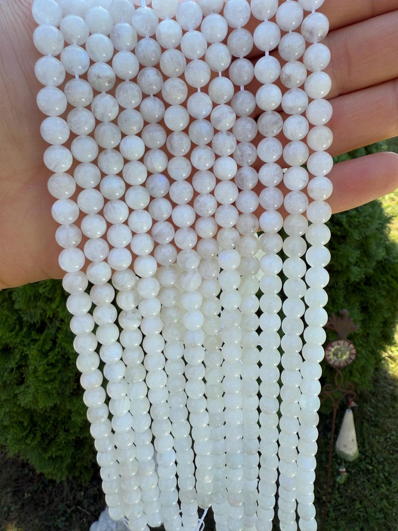 May include: Strands of white, round gemstone beads. The beads have a smooth, polished surface and are strung together on a white cord. The beads are a milky white color with subtle variations in tone. The background is a blurred green.