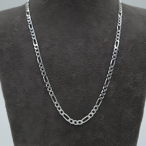 Può includere: Una collana a catena Figaro argentata esposta su un manichino grigio scuro. La collana presenta un motivo di tre piccoli anelli circolari seguiti da un anello ovale allungato, creando un design ripetitivo. La catena è realizzata in metallo lucido e riflettente.