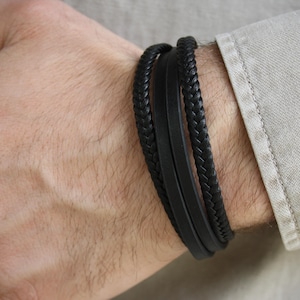 Könnte beinhalten: Schwarzes Lederarmband mit drei Strängen. Ein Strang ist geflochten, die anderen beiden sind glatt. Das Armband wird an einem Handgelenk neben einem hellen Hemd getragen.