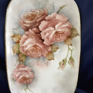 May include: A white porcelain serving dish with a gold rim features a hand-painted design of pink roses and green leaves. The dish measures 22 cm long by 16 cm wide.