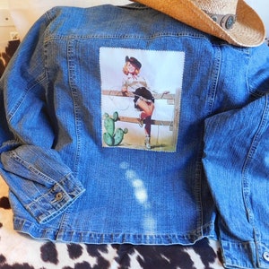 May include: A blue denim jacket with a patch on the back featuring a woman in a cowboy hat and skirt standing on a fence with a cactus in the background.