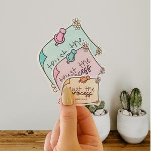 May include: Three pastel-colored stickers with the phrase "Trust the Process" written in black script. The stickers are layered on top of each other and have a whimsical, hand-drawn style with small flowers and push pins.