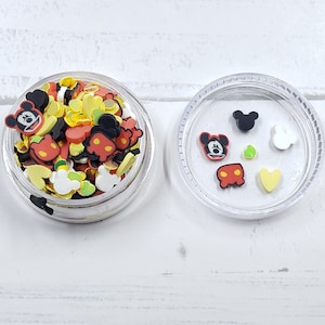 May include: A clear plastic container filled with colorful sprinkles and small plastic charms featuring Mickey Mouse. The container is open and the lid is next to it. The lid has a few of the charms inside.