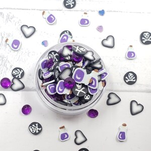 May include: A clear jar filled with purple, black, and white resin charms. The charms are shaped like hearts, bottles, and skulls. The jar is surrounded by loose charms on a white surface.