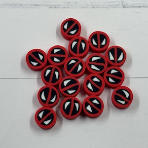 Op de afbeelding: Een verzameling kleine, ronde, rode en zwarte kralen met een wit grafisch ontwerp. De kralen zijn verspreid op een wit oppervlak. Het ontwerp heeft een witte oogvorm met een zwarte omtrek.