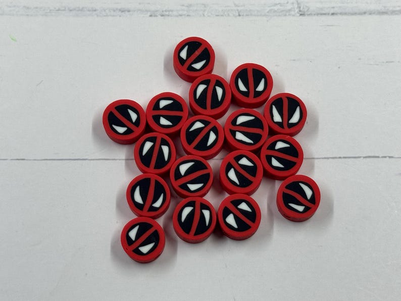 Op de afbeelding: Een verzameling kleine, ronde, rode en zwarte kralen met een wit grafisch ontwerp. De kralen zijn verspreid op een wit oppervlak. Het ontwerp heeft een witte oogvorm met een zwarte omtrek.