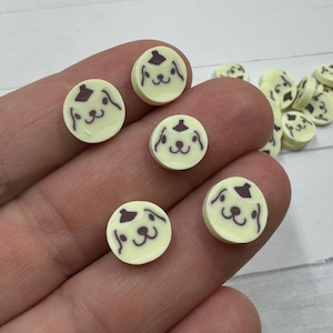 May include: A collection of small, round, light yellow embellishments with a dog face design in dark brown. Several are held in a hand, with more scattered in the background. The dog face has a hat.