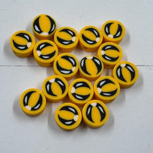 Op de afbeelding: Een verzameling gele, ronde kralen met een zwart-wit ontwerp. De kralen hebben een gele basis met een zwart gebogen ontwerp en een witte accent. De kralen zijn verspreid op een wit oppervlak.