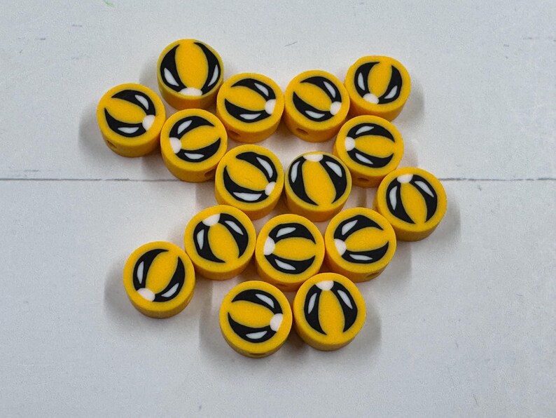 Op de afbeelding: Een verzameling gele, ronde kralen met een zwart-wit ontwerp. De kralen hebben een gele basis met een zwart gebogen ontwerp en een witte accent. De kralen zijn verspreid op een wit oppervlak.