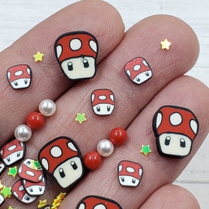 May include: A close-up of red and white mushroom-shaped charms with black eyes and a white spot on top. The charms are scattered on a hand with white and yellow star-shaped sprinkles.