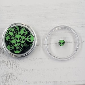 May include: Two clear plastic containers, one filled with green alien face stickers and the other with a single green alien face sticker.